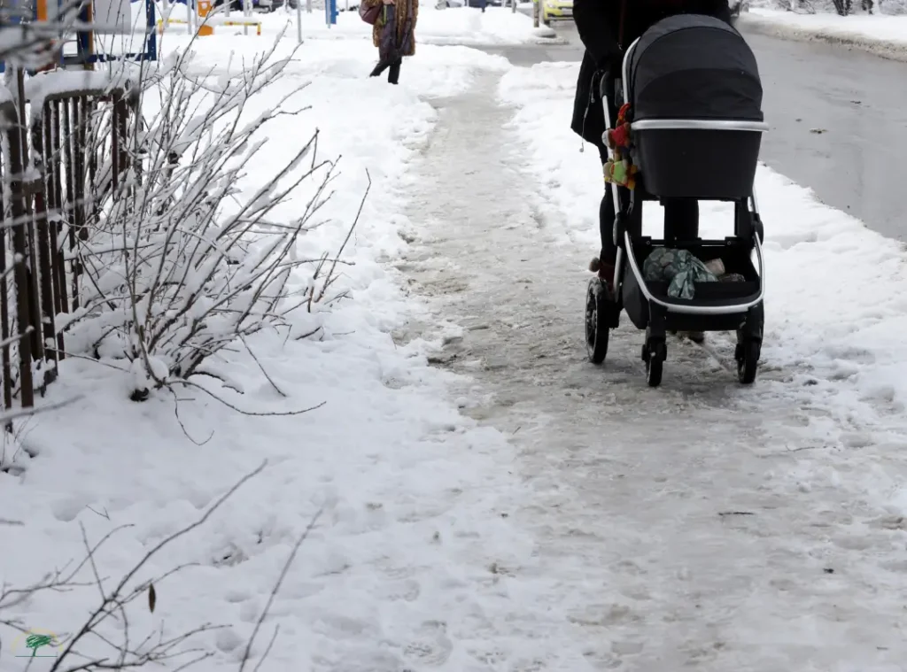A person pushes a baby stroller along an icy, partially cleared sidewalk bordered by snow, illustrating the risks of refreezing and black ice after daytime melt in coastal Massachusetts towns.